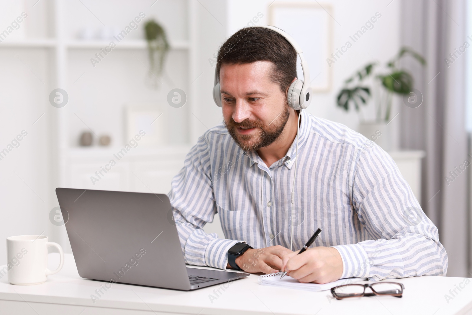 Photo of Interpreter in headphones taking notes while having video chat via laptop at white table indoors