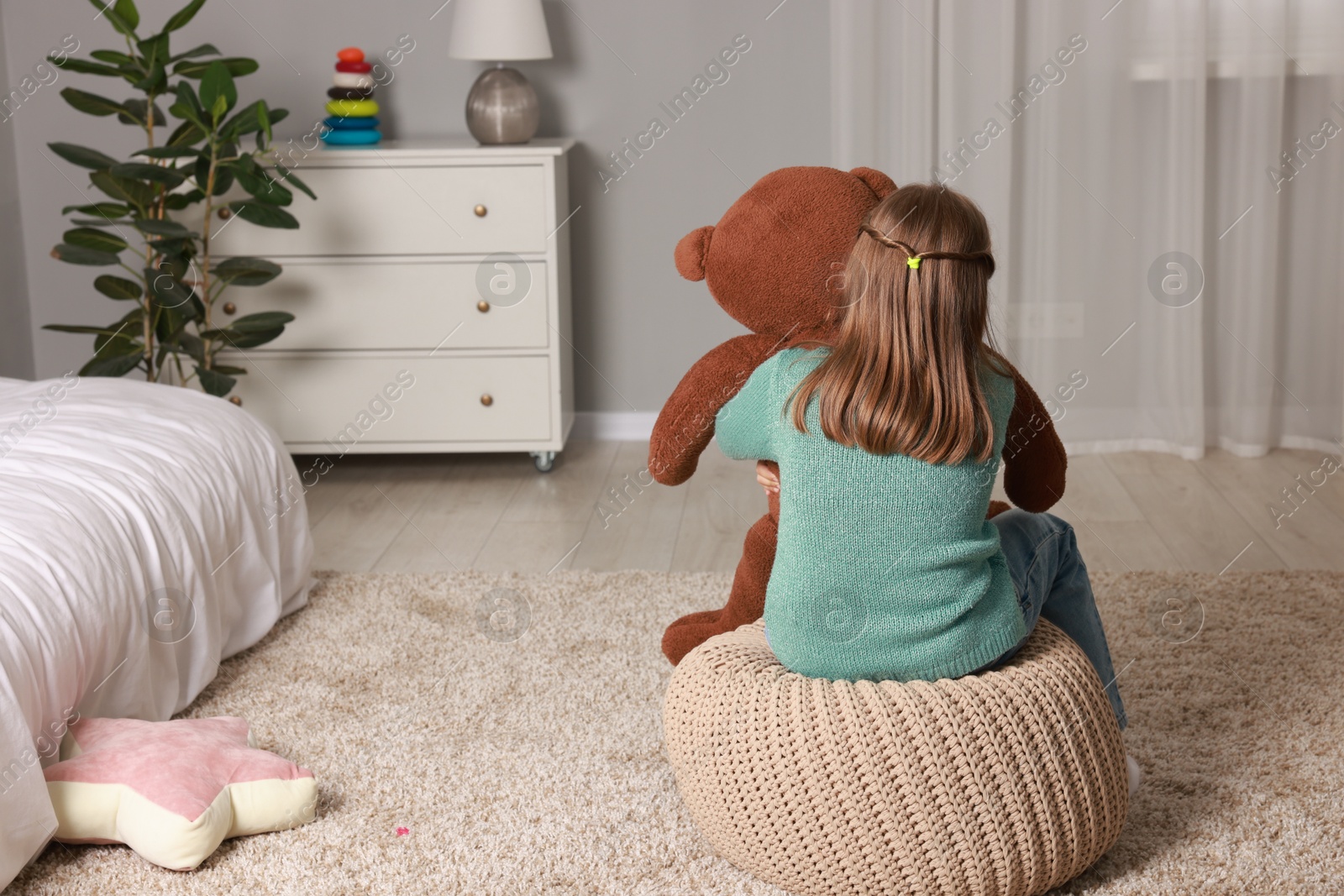 Photo of Autism concept. Lonely little girl with teddy bear on pouf at home, back view