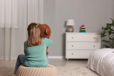 Autism concept. Lonely little girl with teddy bear on pouf at home, back view