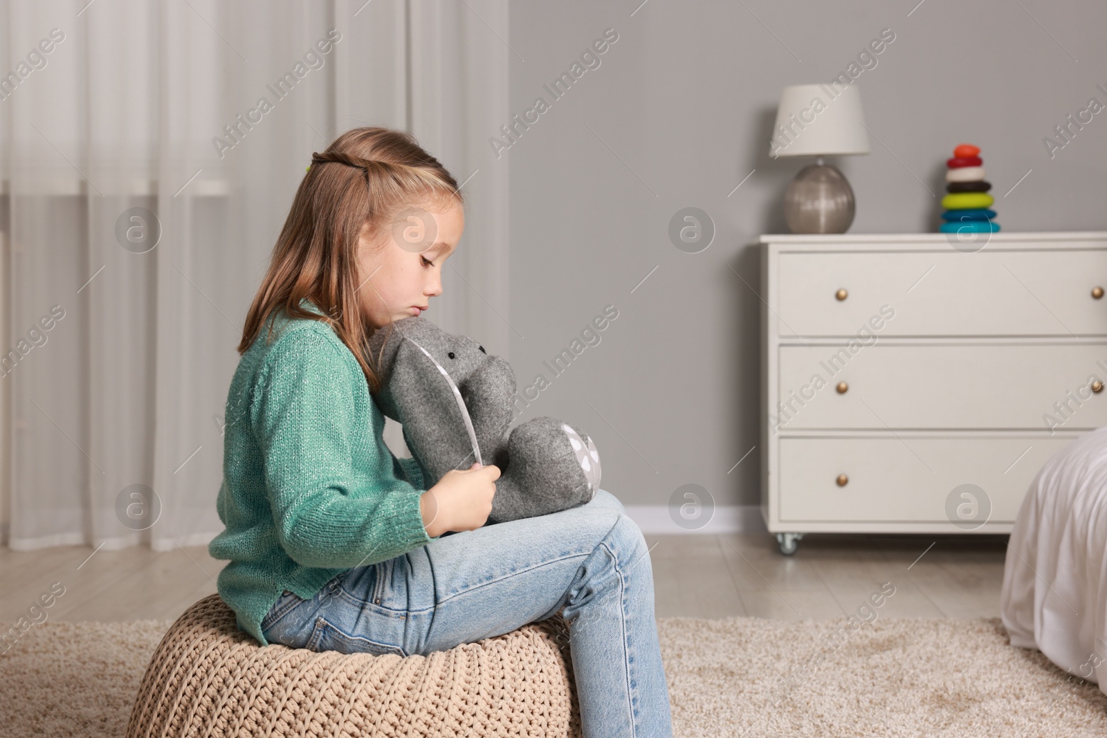 Photo of Autism concept. Lonely little girl with toy bunny on pouf at home