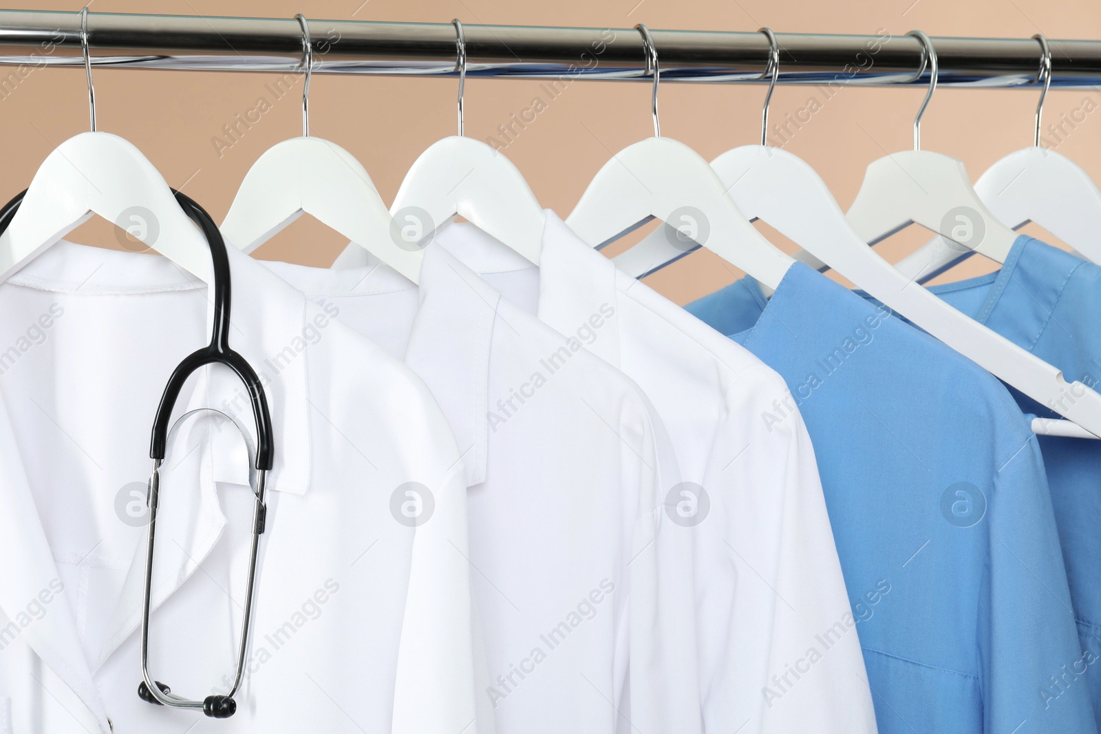 Photo of Different medical workers' uniforms and stethoscope on clothing rack against beige background, closeup