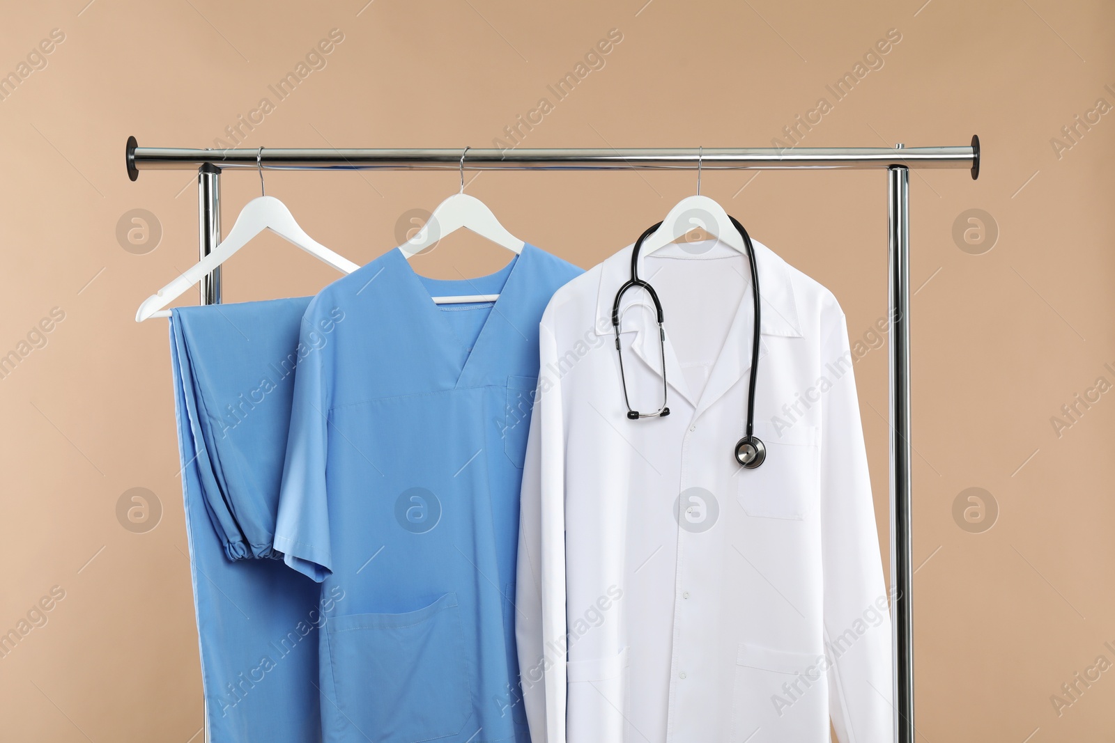 Photo of Different medical workers' uniforms and stethoscope on clothing rack against beige background