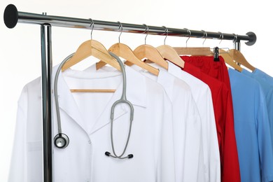 Photo of Different medical workers' uniforms and stethoscope on clothing rack against white background