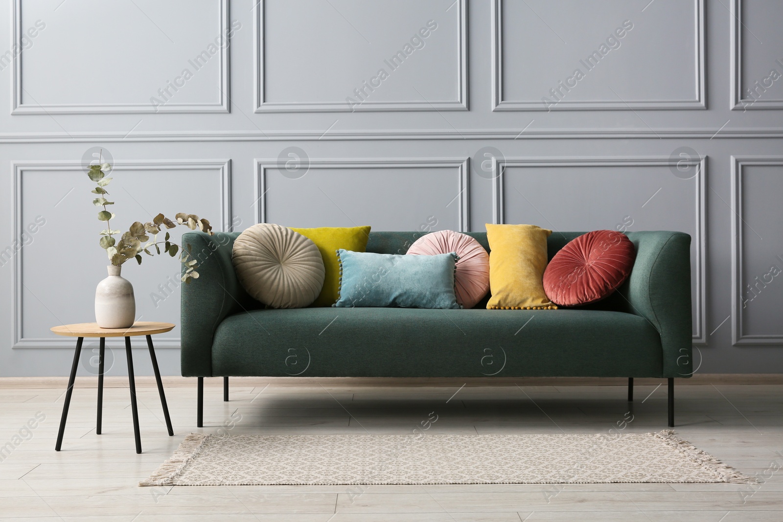 Photo of Stylish sofa with cushions and eucalyptus branches in vase on side table near grey wall