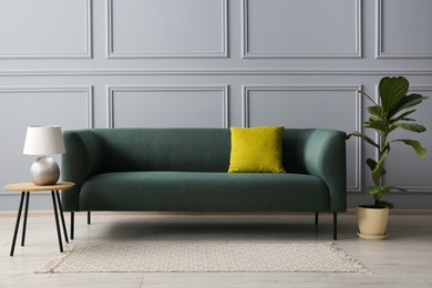 Stylish sofa with cushion, lamp on side table and green plant near grey wall