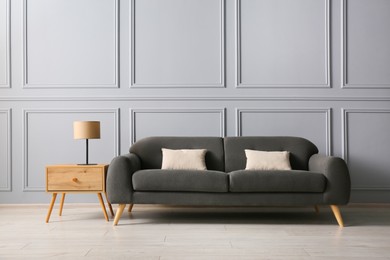 Stylish sofa with cushions and lamp on side table near grey wall