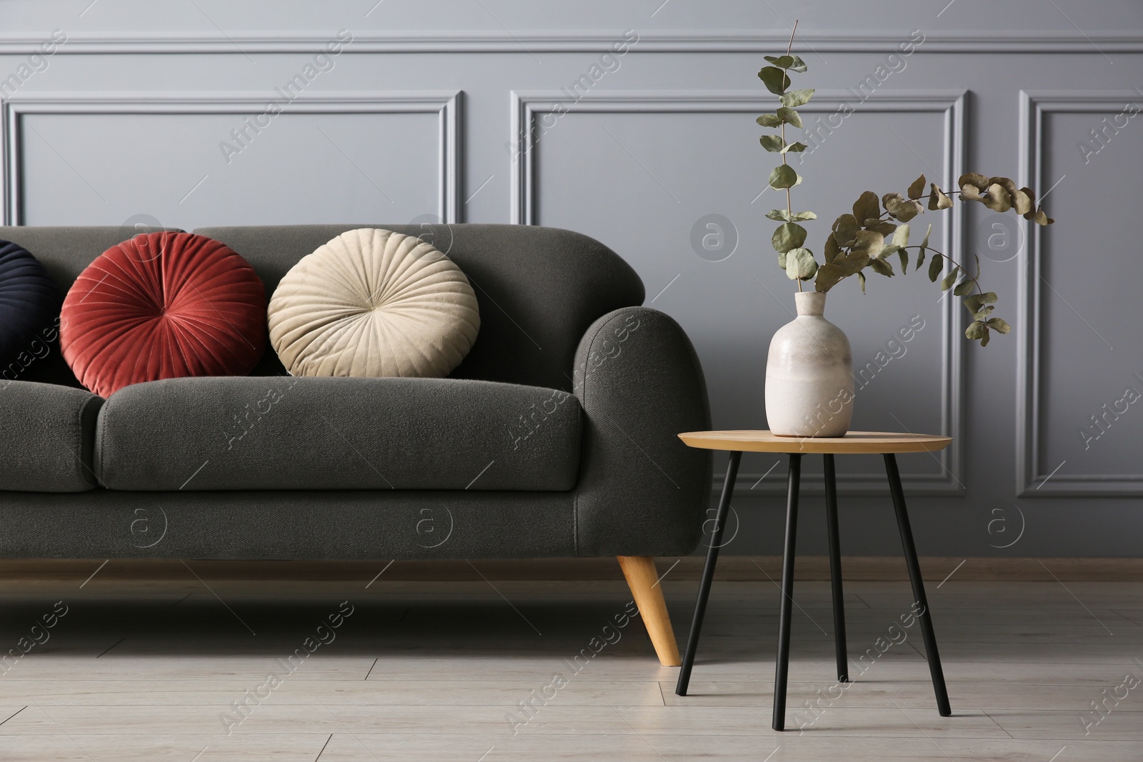 Photo of Stylish sofa with cushions and eucalyptus branches in vase on coffee table near grey wall