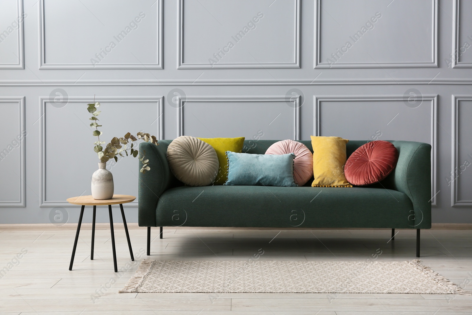 Photo of Stylish sofa with cushions and eucalyptus branches in vase on side table near grey wall