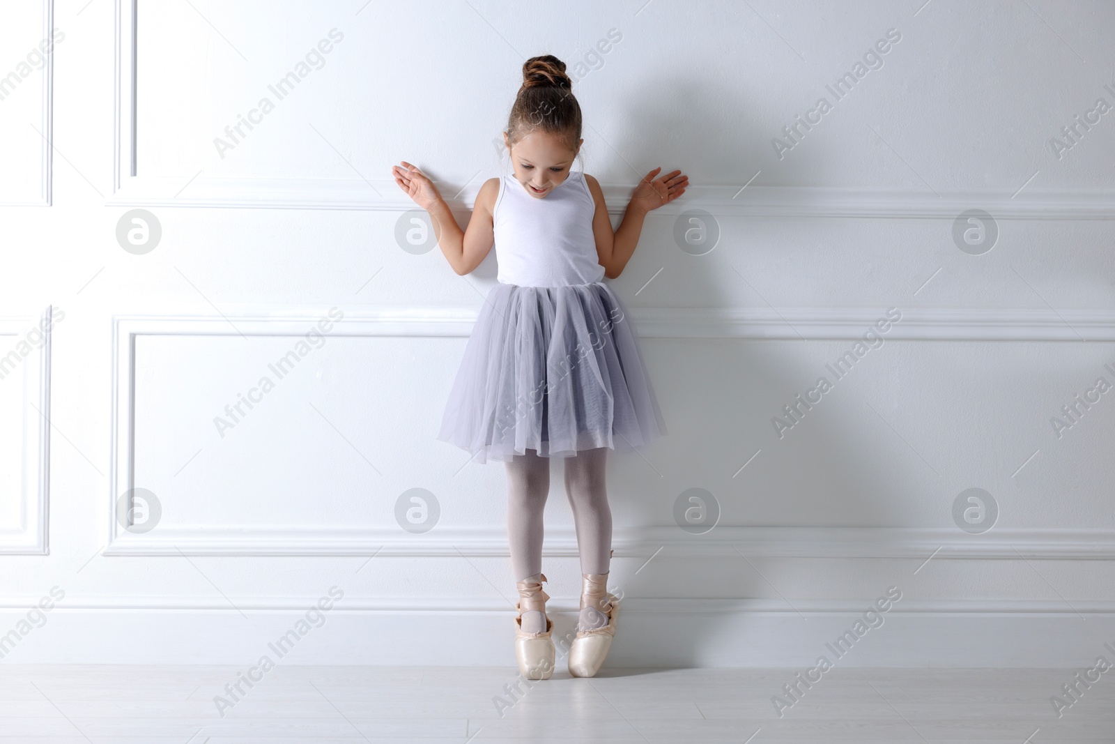 Photo of Little ballerina practicing dance moves near white wall indoors