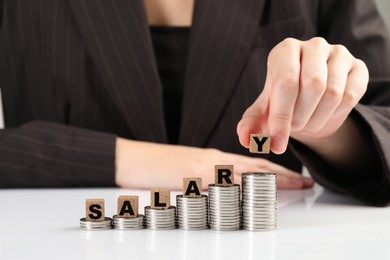 Photo of Woman putting wooden cubes with word Salary and stacks of coins on white table, closeup