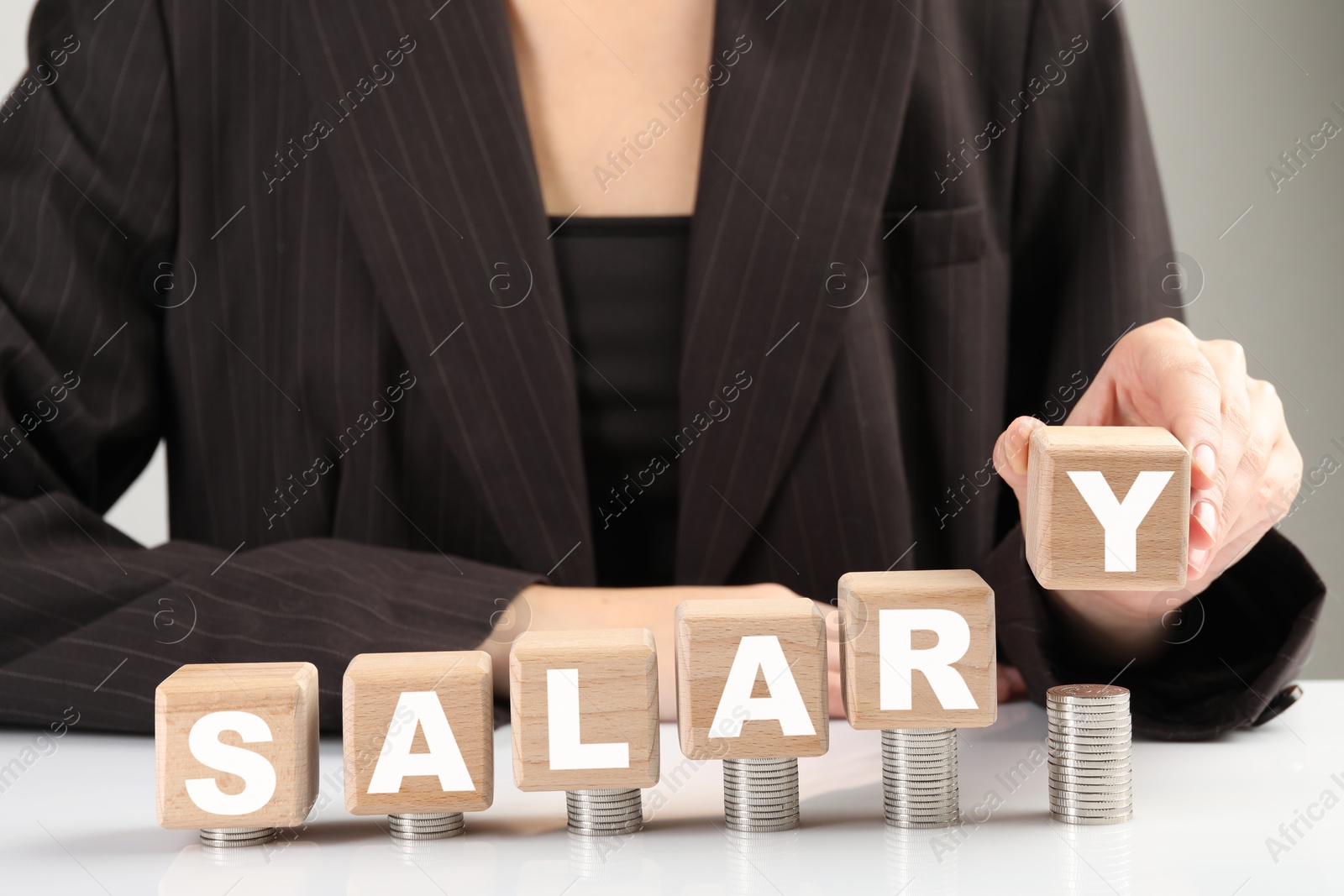 Photo of Woman putting wooden cubes with word Salary and stacks of coins on white table, closeup