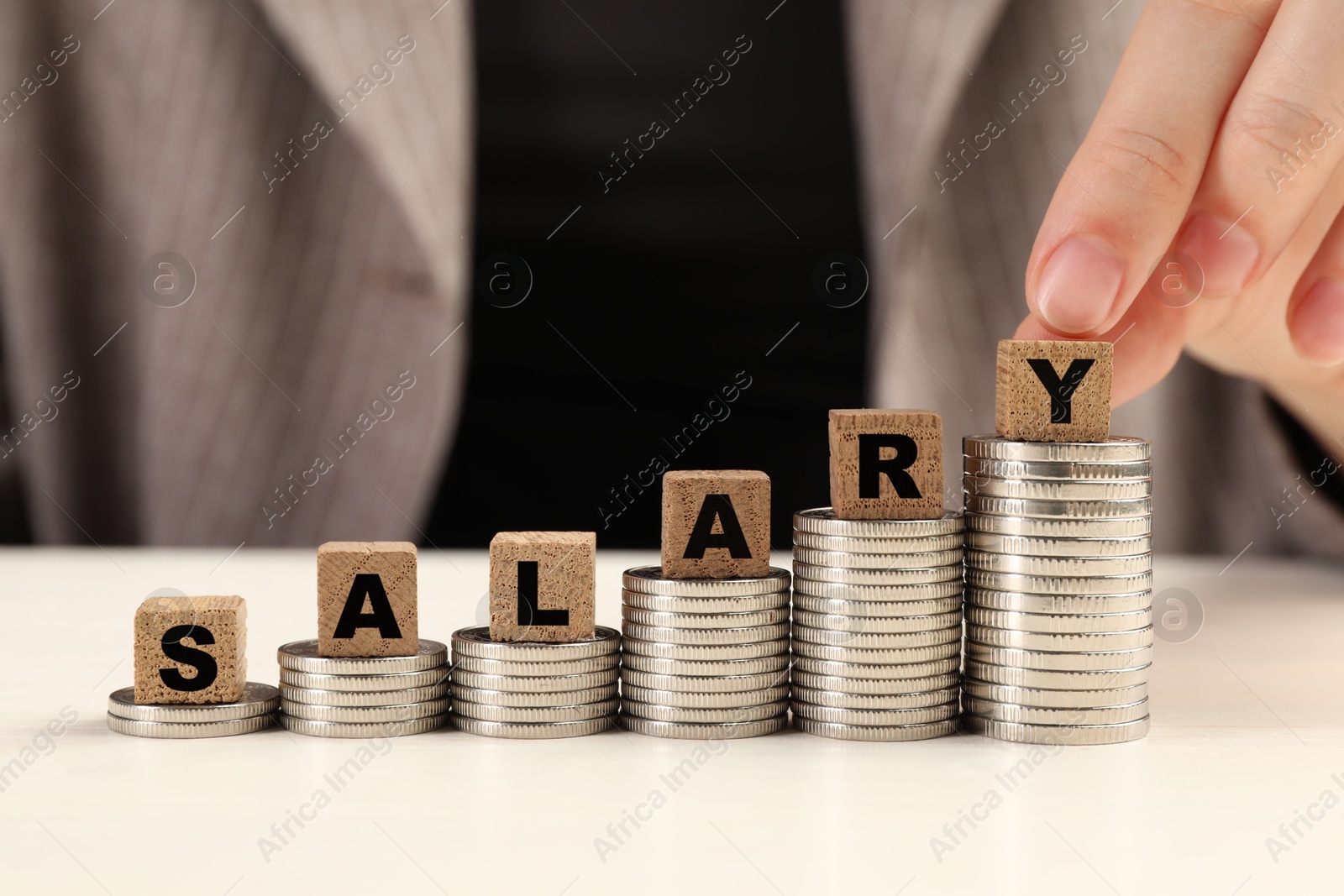 Photo of Woman putting wooden cubes with word Salary and stacks of coins on white table, closeup