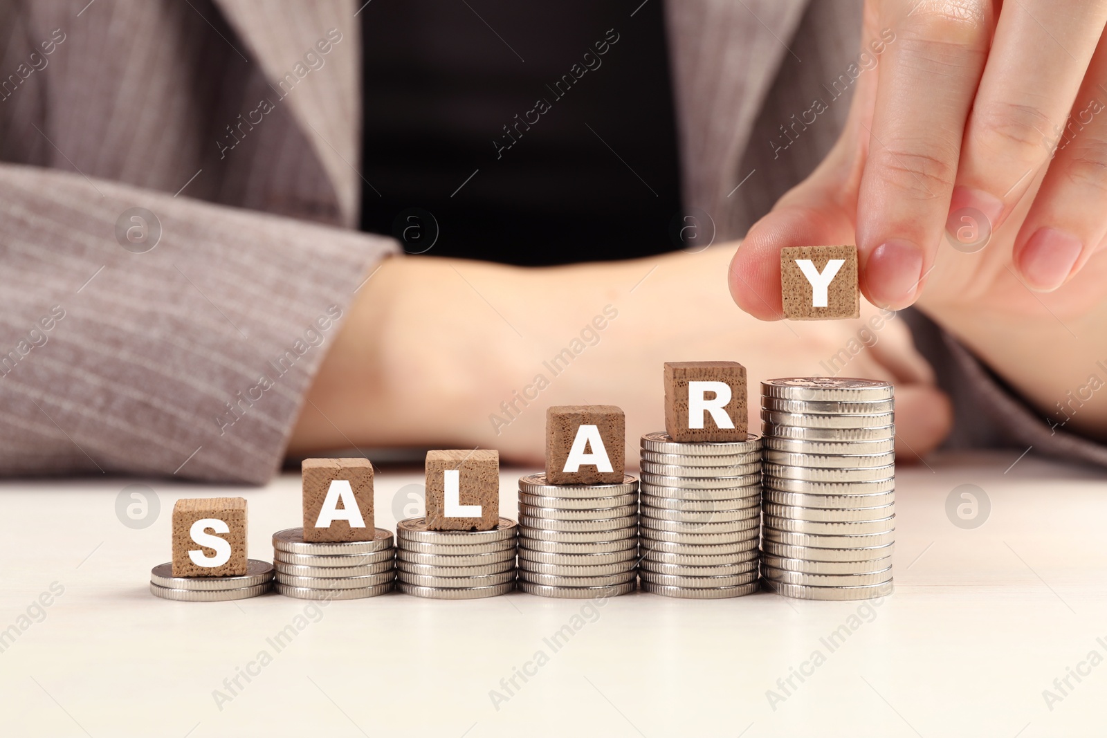 Photo of Woman putting wooden cubes with word Salary and stacks of coins on white table, closeup