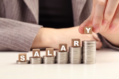 Photo of Woman putting wooden cubes with word Salary and stacks of coins on white table, closeup