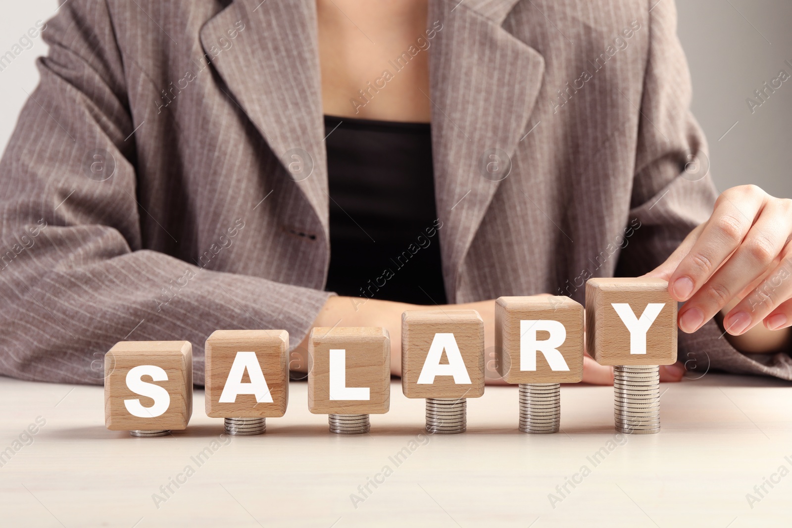 Photo of Woman putting wooden cubes with word Salary and stacks of coins on white table, closeup