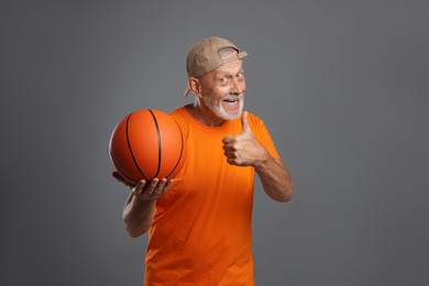 Portrait of happy senior man with basketball ball showing thumbs up on grey background