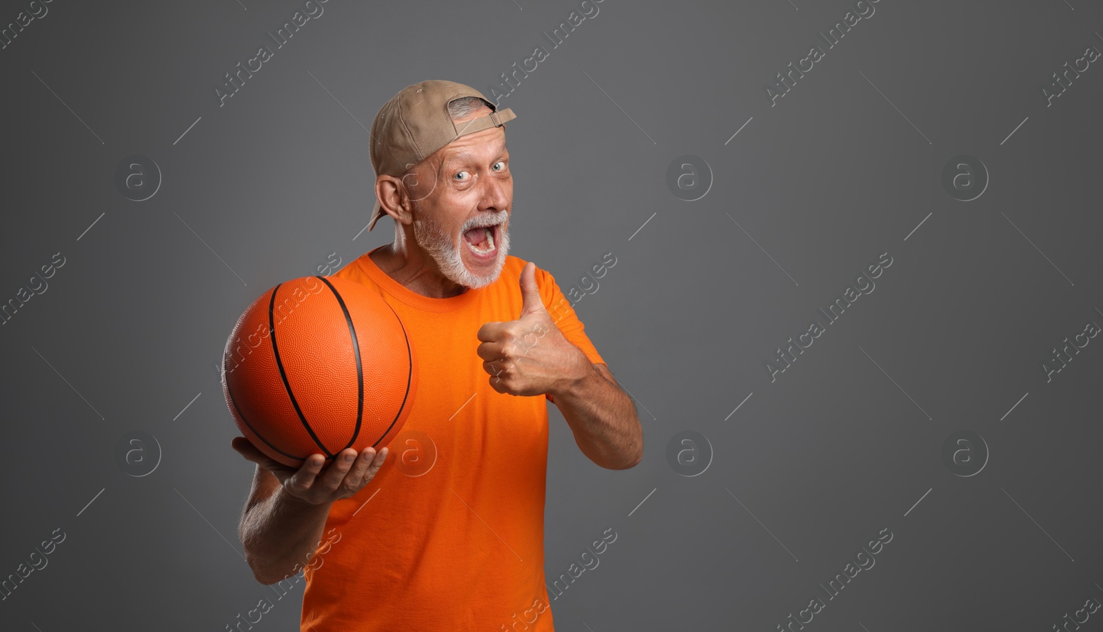 Photo of Portrait of emotional senior man with basketball ball showing thumbs up on grey background. Space for text