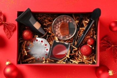 Photo of Christmas gift box with cosmetic products and festive decor on red background, top view