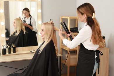 Photo of Hairdresser curling woman's hair with flat iron in salon