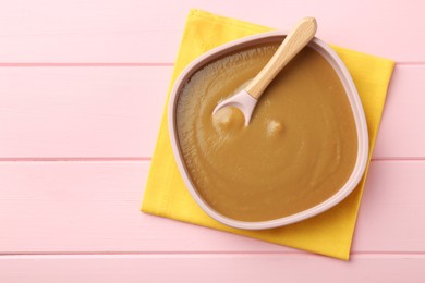 Photo of Delicious baby food and spoon in bowl on pink wooden table, top view