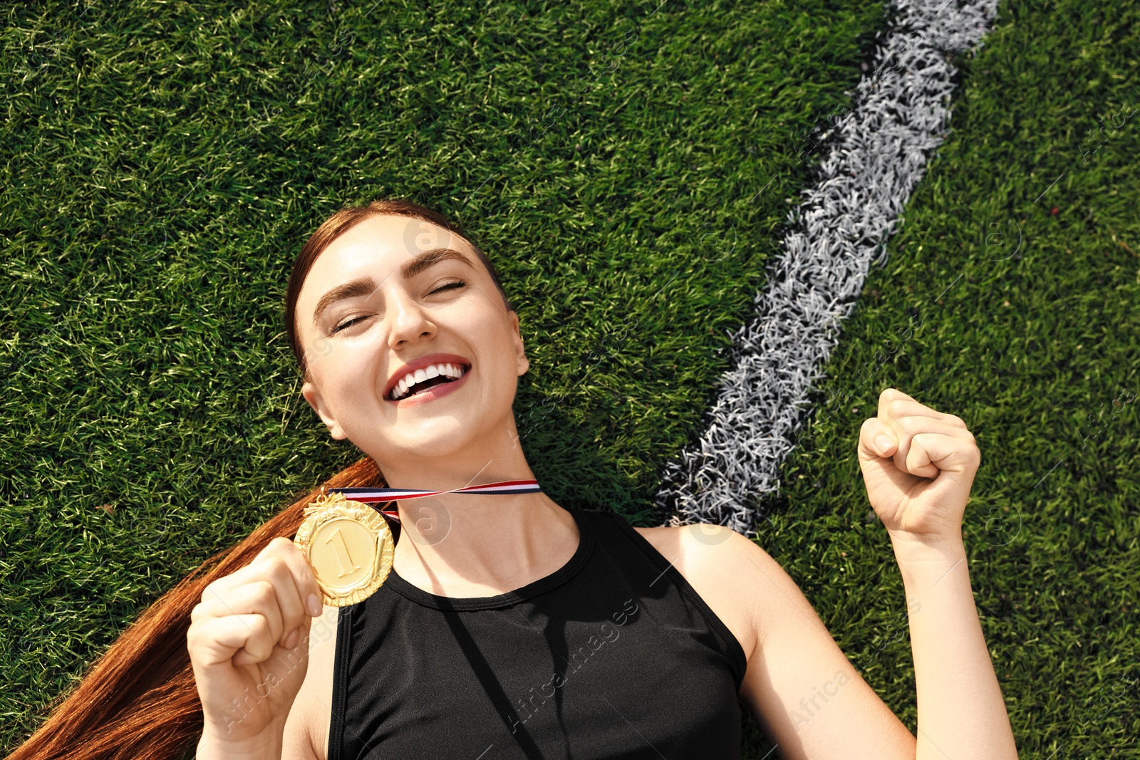 Photo of Happy winner with golden medal lying on green grass at stadium, top view