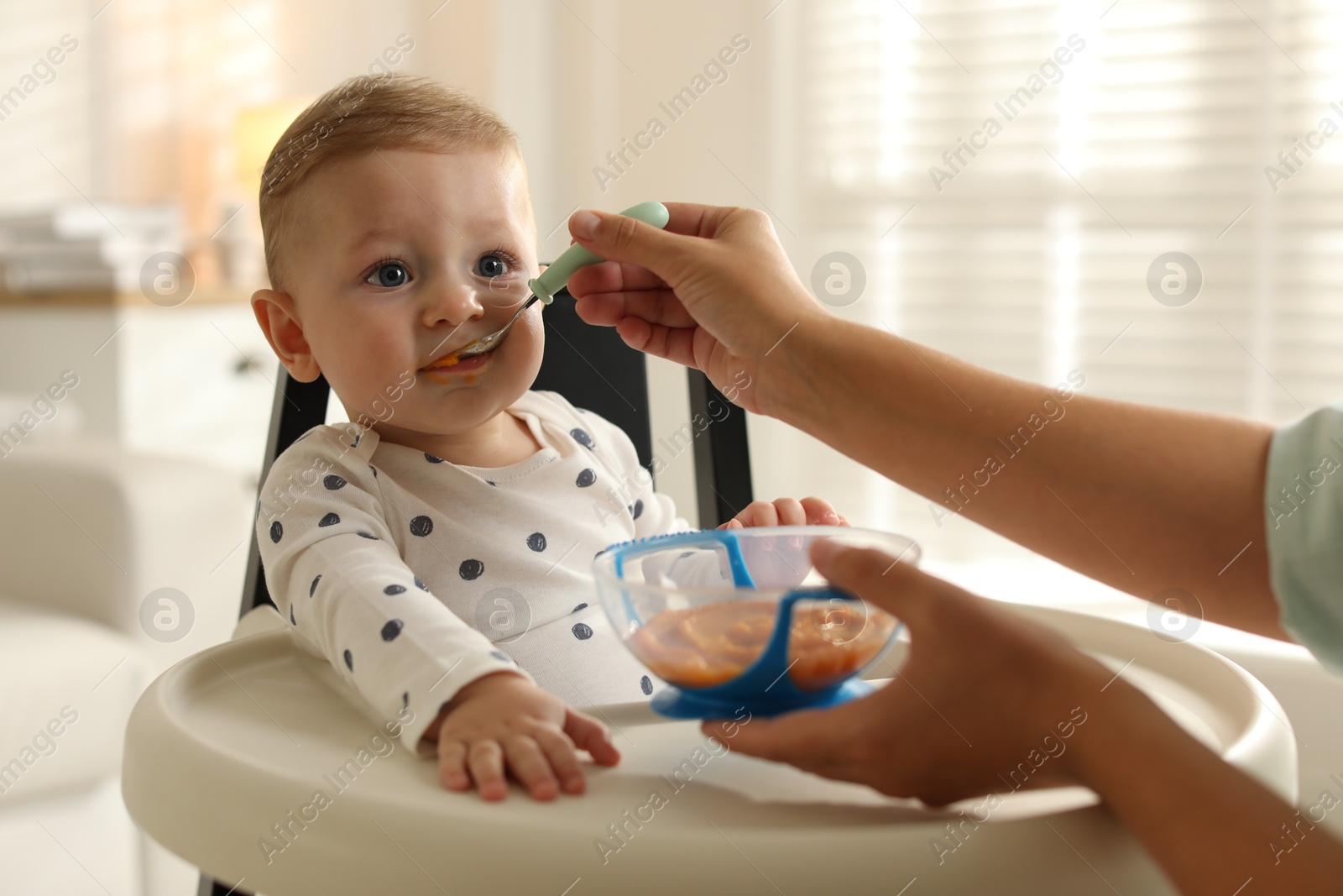 Photo of Mother feeding her cute little baby in high chair at home, closeup