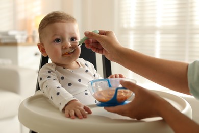 Photo of Mother feeding her cute little baby in high chair at home, closeup