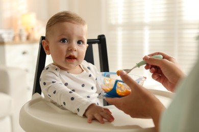 Mother feeding her cute little baby in high chair at home, closeup