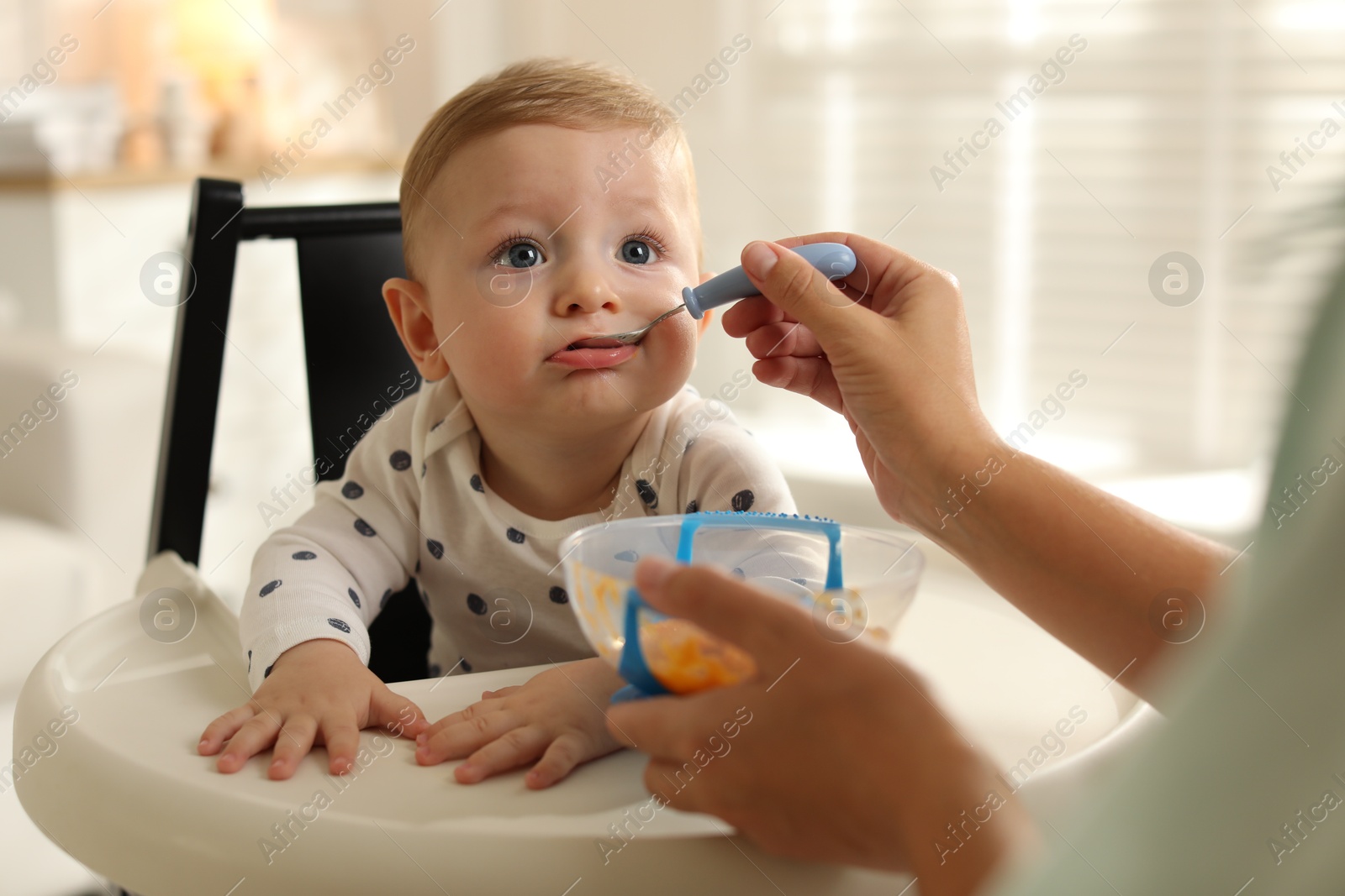 Photo of Mother feeding her cute little baby in high chair at home, closeup