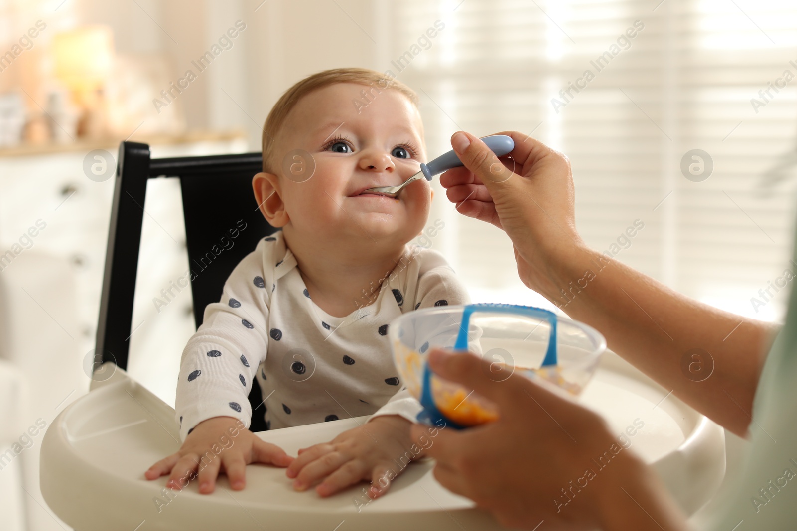 Photo of Mother feeding her cute little baby in high chair at home, closeup