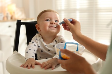 Mother feeding her cute little baby in high chair at home, closeup