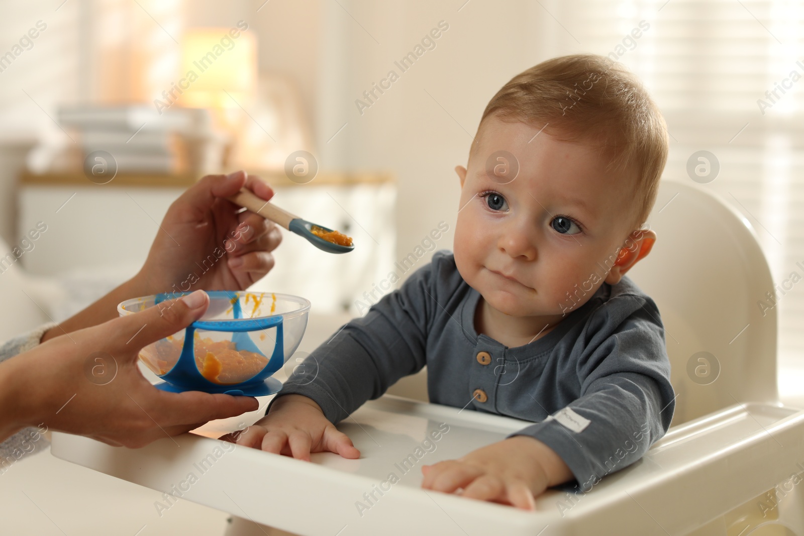 Photo of Mother feeding her cute little baby in high chair at home, closeup