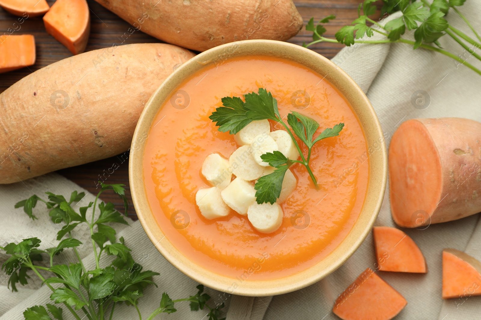 Photo of Delicious sweet potato soup with croutons in bowl, fresh vegetables and parsley on wooden table, flat lay