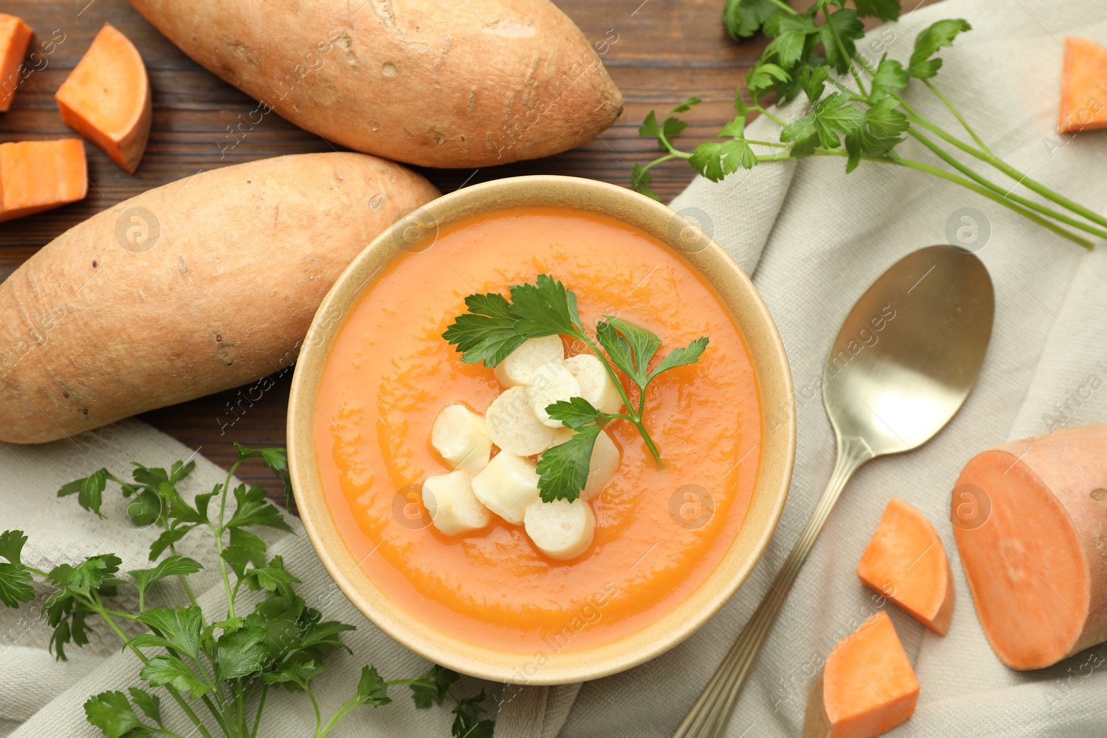 Photo of Delicious sweet potato soup with croutons in bowl, fresh vegetables and parsley on wooden table, flat lay