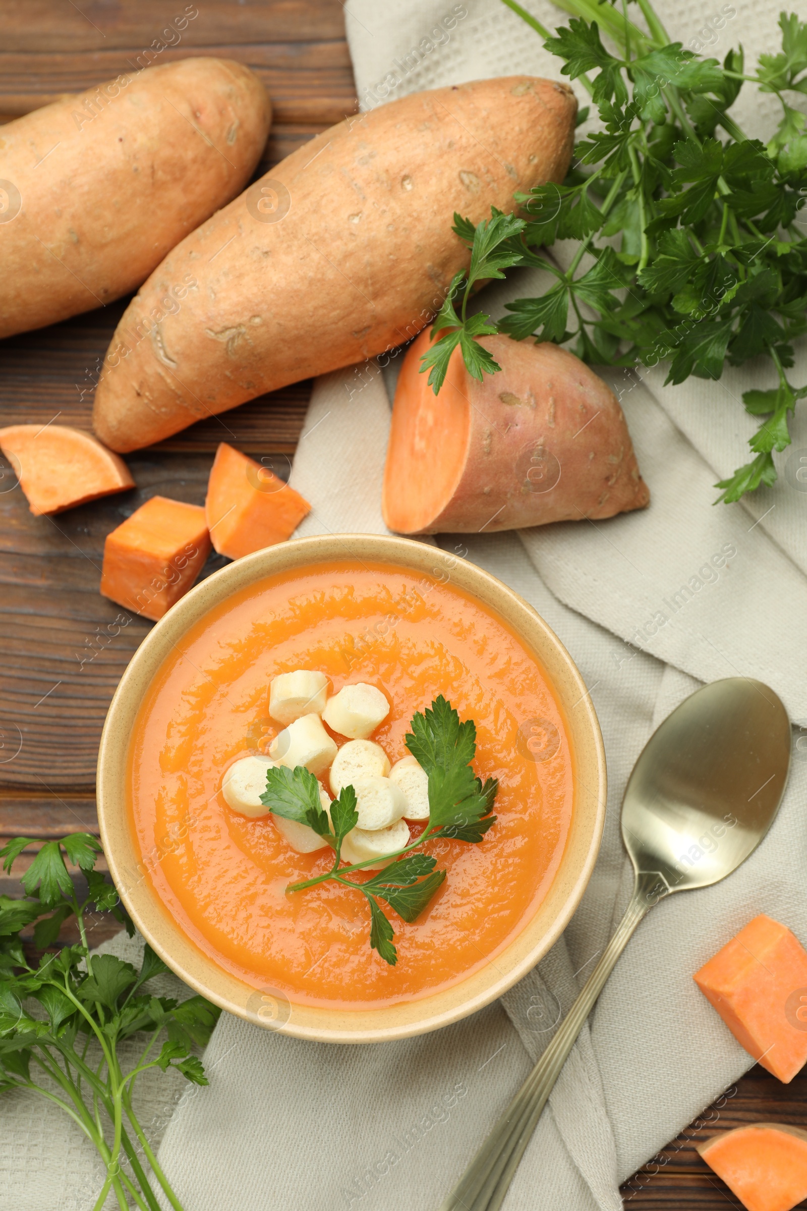 Photo of Delicious sweet potato soup with croutons in bowl, fresh vegetables and parsley on wooden table, flat lay