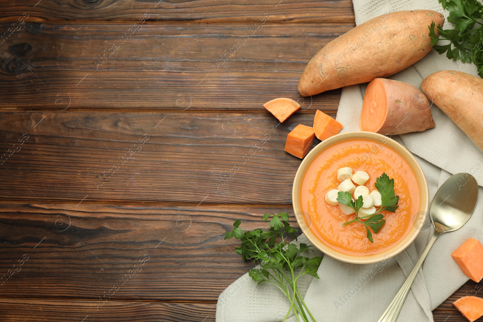 Photo of Delicious sweet potato soup with croutons in bowl, fresh vegetables and parsley on wooden table, flat lay. Space for text