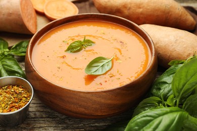 Photo of Delicious sweet potato soup with spices in bowl, fresh vegetables and basil on wooden table, closeup