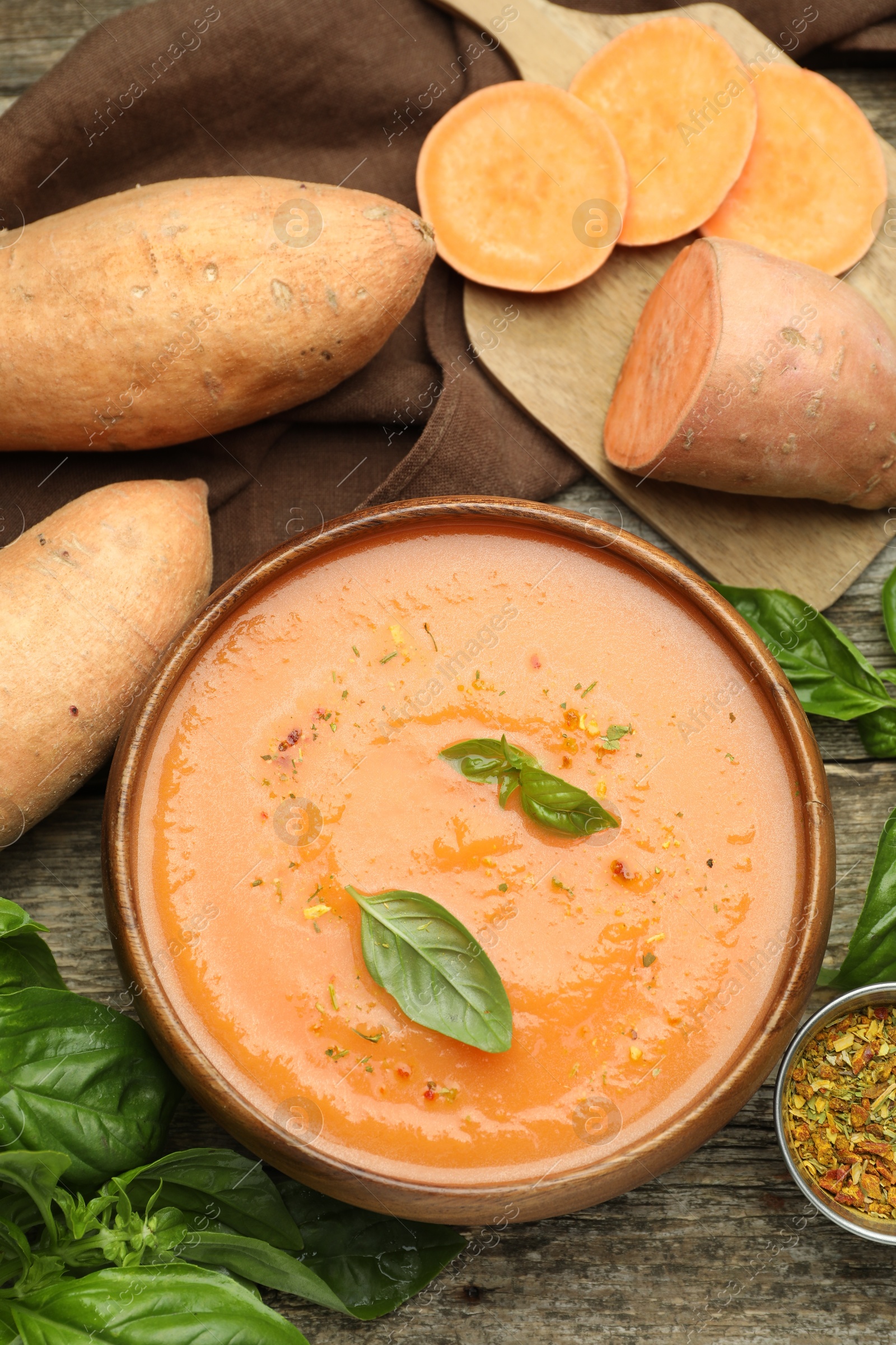 Photo of Delicious sweet potato soup with spices in bowl, fresh vegetables and basil on wooden table, flat lay