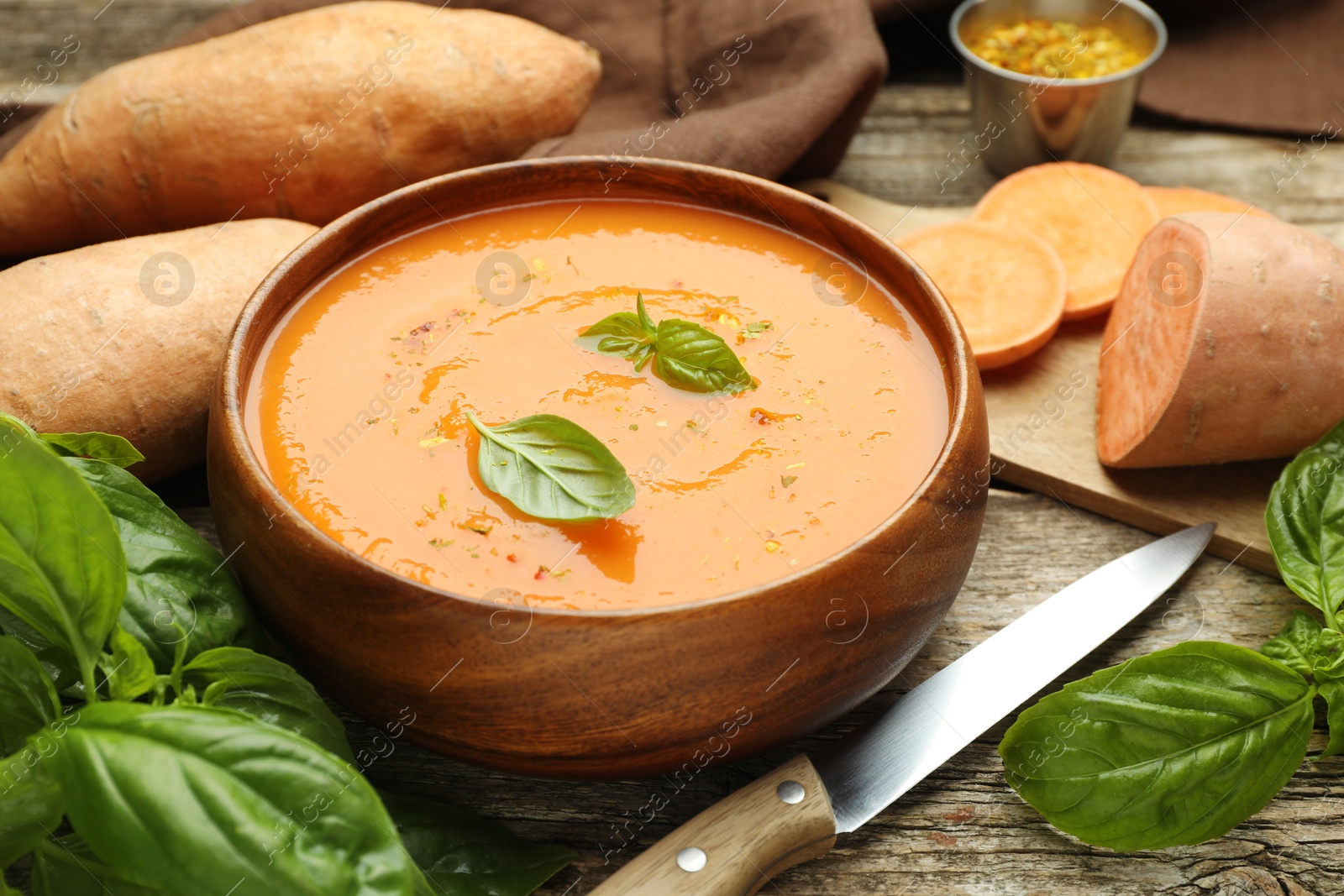 Photo of Delicious sweet potato soup, fresh vegetables, basil and knife on wooden table, closeup