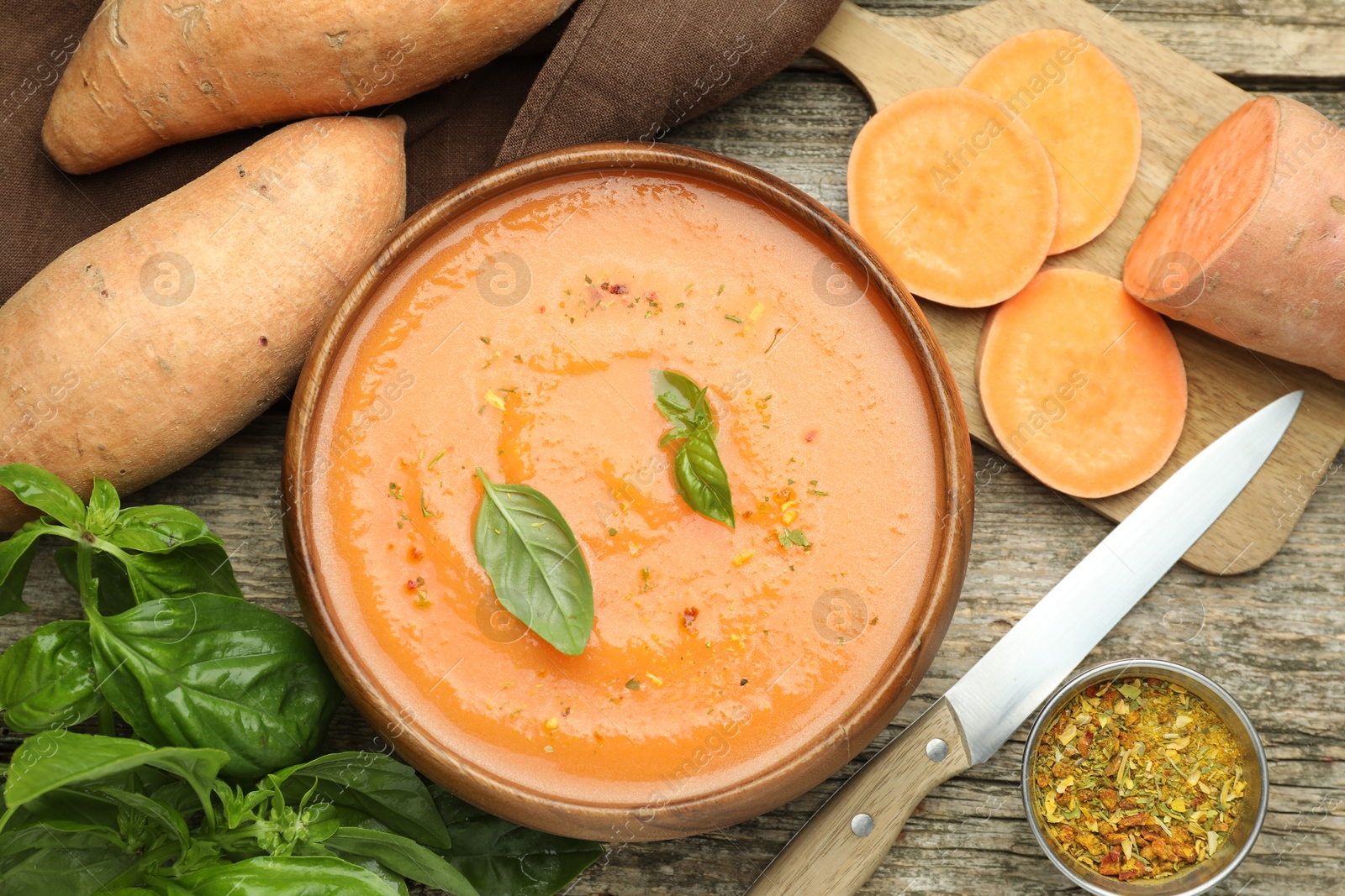 Delicious sweet potato soup with spices in bowl, fresh vegetables, basil and knife on wooden table, flat lay Photo of Delicious sweet potato soup with spices in bowl, fresh vegetables, basil and knife on wooden table, flat lay