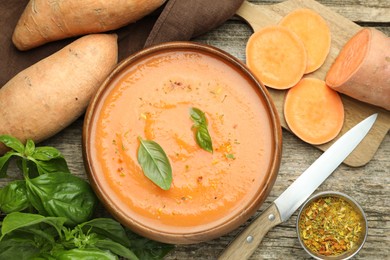 Photo of Delicious sweet potato soup with spices in bowl, fresh vegetables, basil and knife on wooden table, flat lay