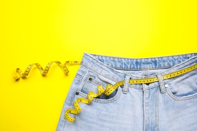 Jeans and measuring tape on yellow background, top view