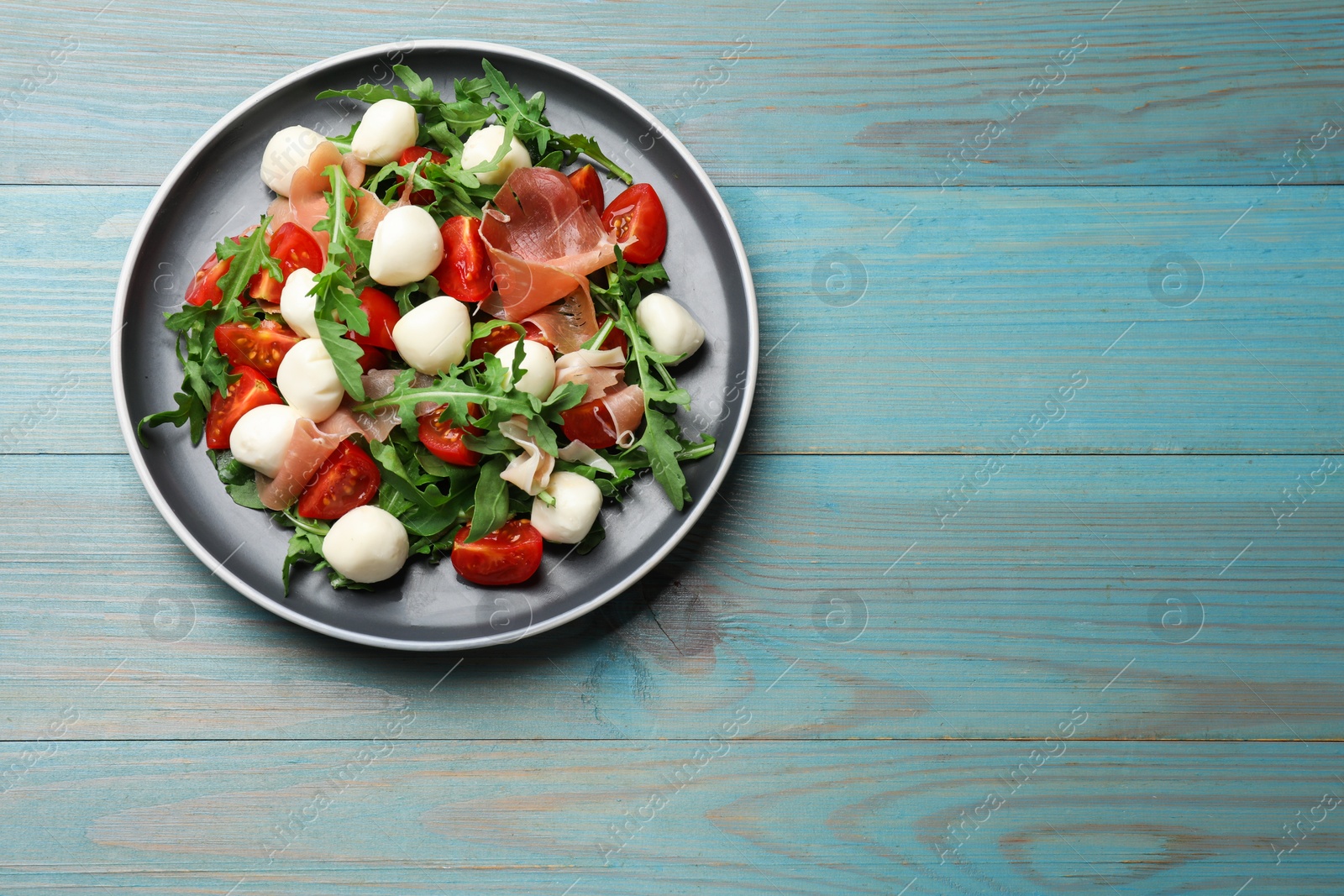 Photo of Tasty salad with arugula, mozzarella cheese, tomatoes and jamon on light blue wooden table, top view. Space for text