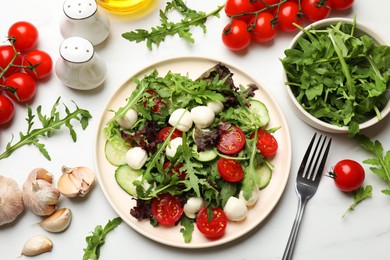 Photo of Tasty salad with arugula, lettuce, mozzarella cheese, vegetables and fork on white table, flat lay