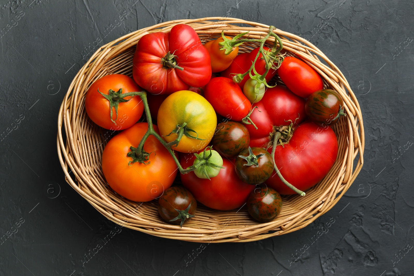 Photo of Different ripe and unripe tomatoes in wicker basket on grey textured table, top view