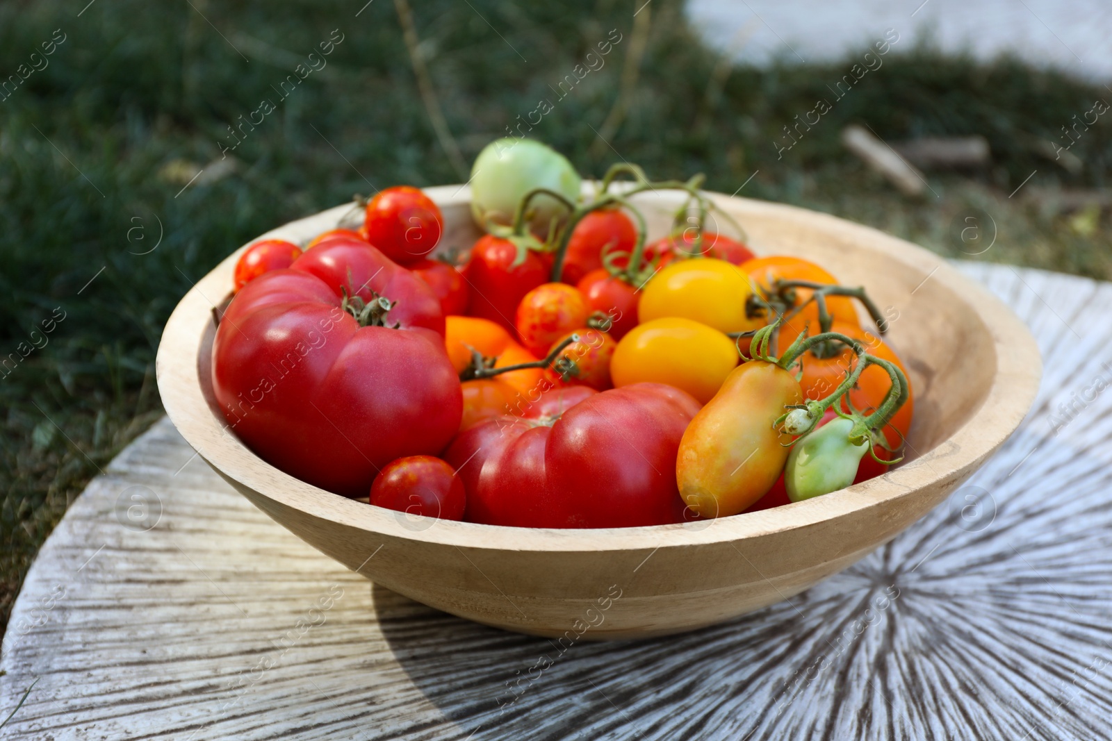 Photo of Different fresh tomatoes in bowl on wooden surface outdoors