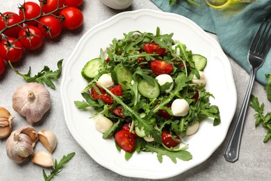 Photo of Tasty salad with arugula, mozzarella, tomatoes and cucumber served on grey textured table, flat lay