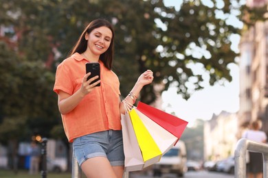 Photo of Happy woman with colorful shopping bags using smartphone outdoors