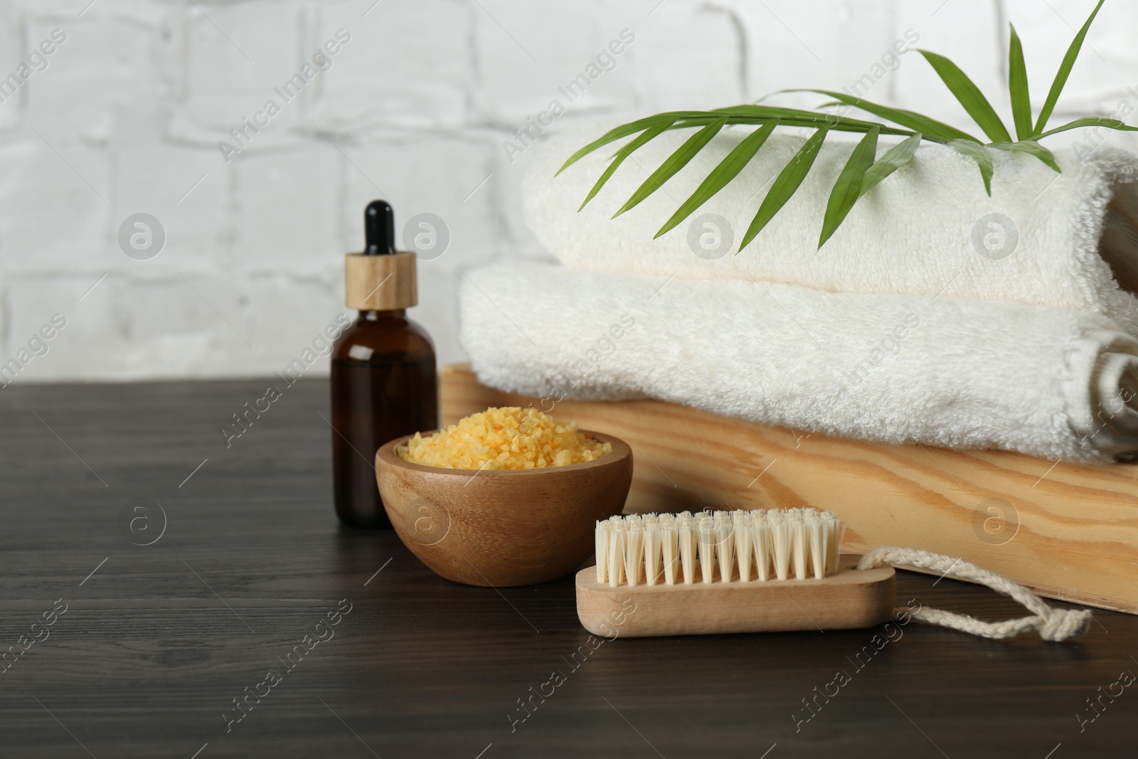 Photo of Spa treatment. Towels, sea salt, brush, bottle of serum and palm leaf on black wooden table, closeup