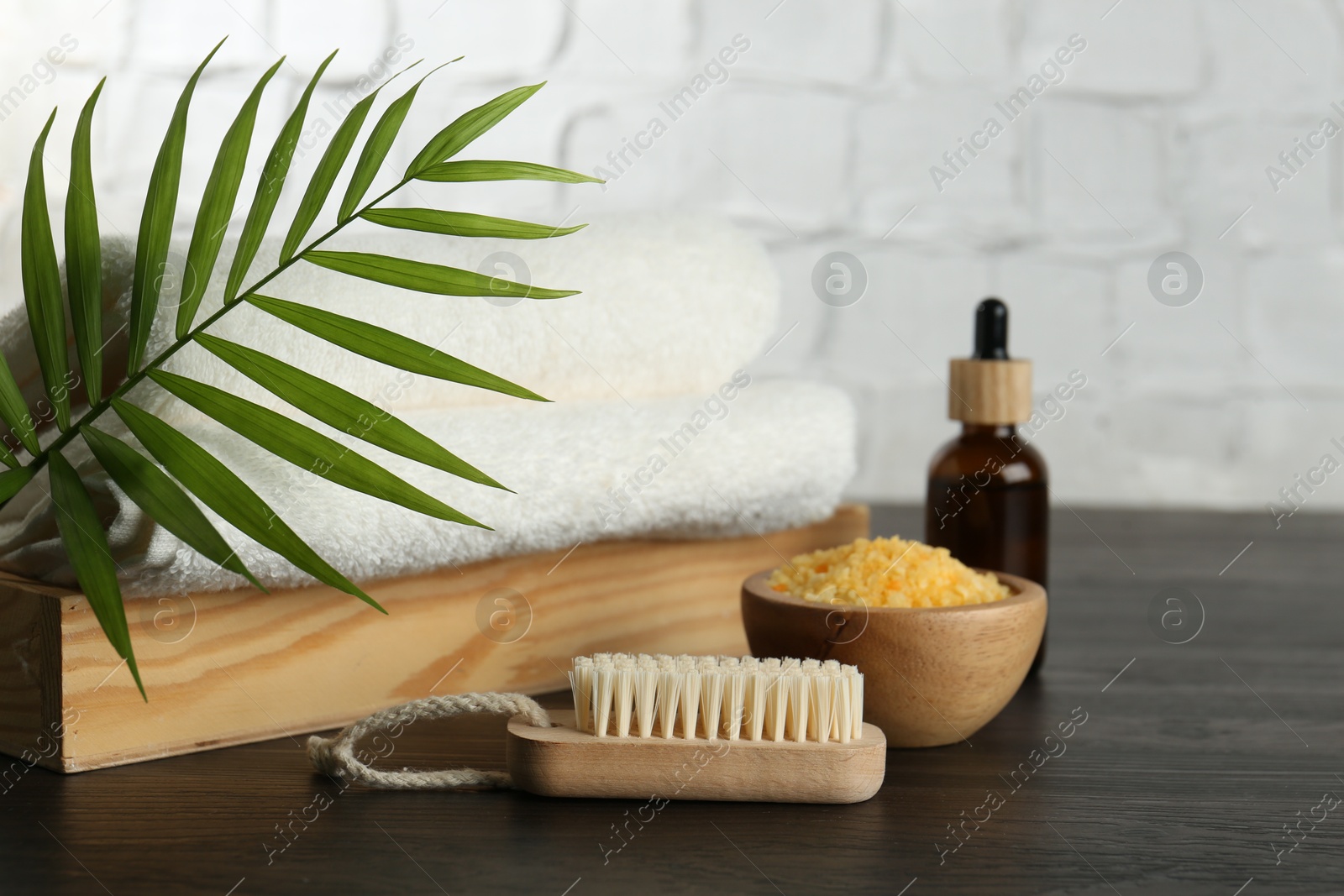 Photo of Spa treatment. Towels, sea salt, brush, bottle of serum and palm leaf on black wooden table, closeup